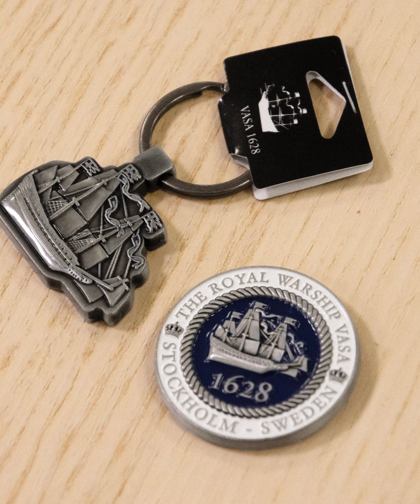 En metallfärgad Vasa örlogsskepp-nyckelring och en rund Vasa 1628-magnet med blå emalj, utlagda på ett träunderlag